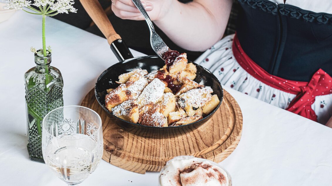 Ein köstliches Gericht in einer schwarzen Pfanne, garniert mit Puderzucker. Im Hintergrund sind ein Glas Wasser und eine Tasse Kaffee zu sehen. | © Anna Ullrich