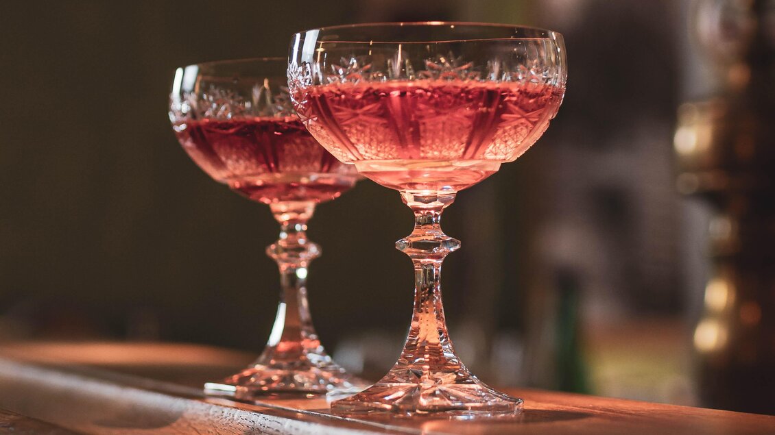 Zwei elegante Cocktailgläser mit rosafarbenem Getränk stehen auf einer Holzbar. Im Hintergrund sind verschwommene Gläser und eine warme, einladende Atmosphäre zu erkennen. | © Anna Ullrich