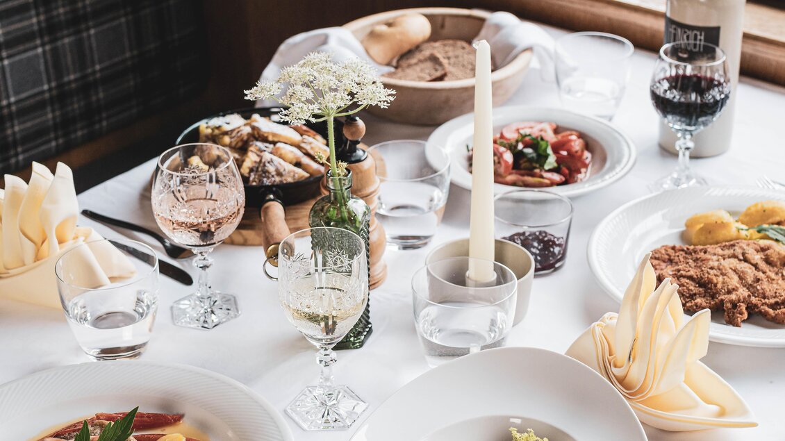 Ein festlich gedeckter Tisch mit verschiedenen Gerichten, darunter Fleisch, Salat und Beilagen. Die Tafel ist mit Gläsern, Kerzen und frischen Blumen dekoriert. | © Anna Ullrich