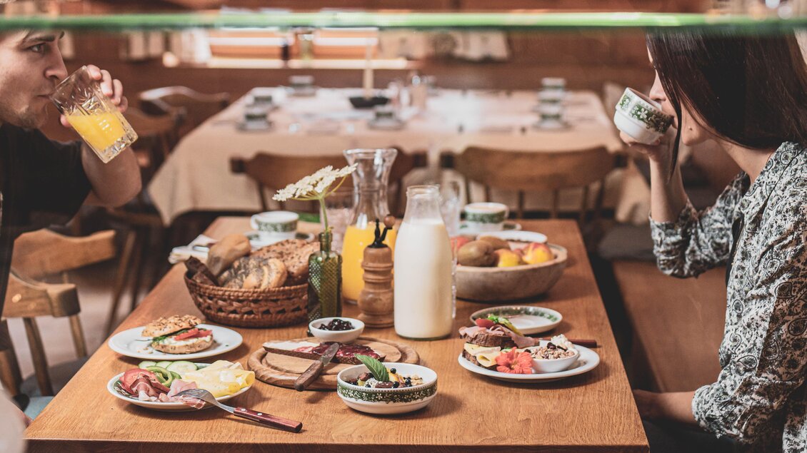 Ein gedeckter Tisch mit verschiedenen Frühstücksgerichten, darunter Brot, Käse, Wurst und Obst. Zwei Personen sitzen entspannt und genießen ihre Getränke. | © Anna Ullrich