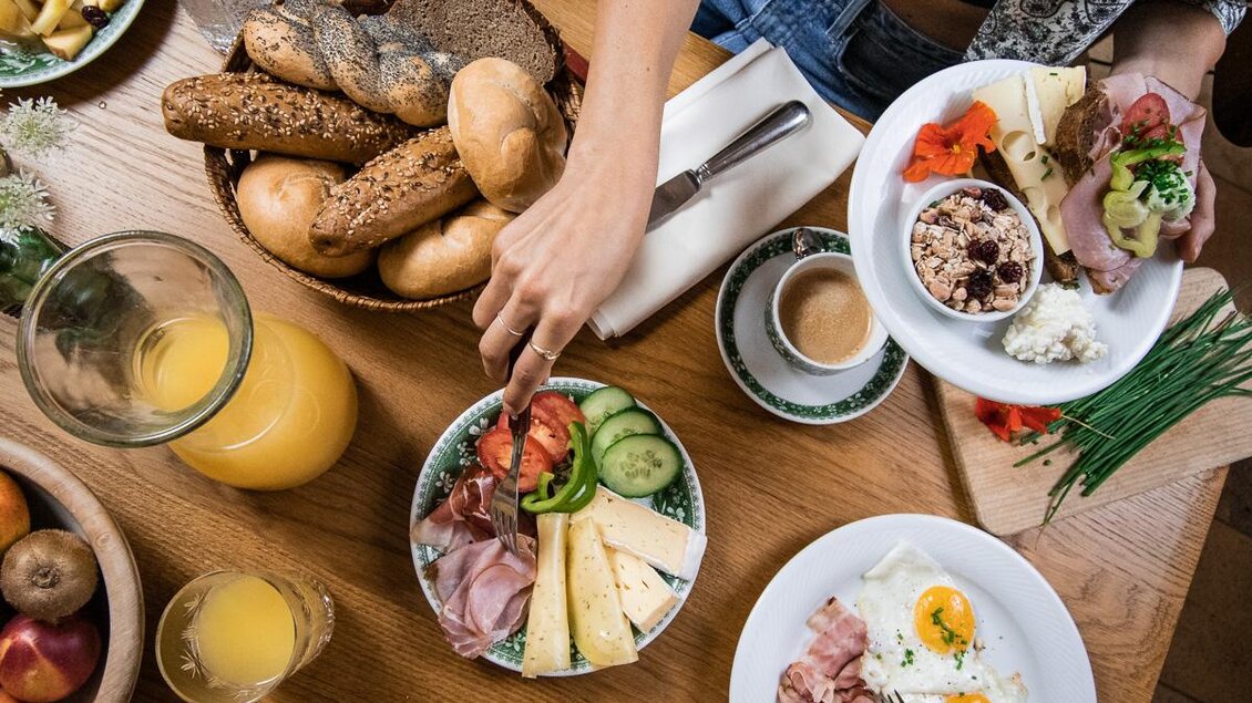 Ein reichhaltiges Frühstück mit frischen Brötchen, Käse, Aufschnitt und verschiedenen Beilagen. Im Vordergrund sind Eier mit Speck und ein Glas Orangensaft zu sehen. | © Anna Ullrich