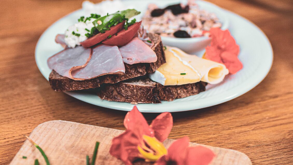 Ein Teller mit belegten Broten, Wurst und Käse, garniert mit frischen Kräutern. Im Hintergrund sind eine Tasse Kaffee und ein weiteres Gericht zu sehen. | © Anna Ullrich
