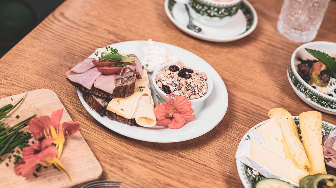 Ein leckerer Frühstücksteller mit Brotscheiben, Wurst, Käse und einer Joghurtschüssel. Dazu gibt es frische Blumen und einen Kaffee. | © Anna Ullrich
