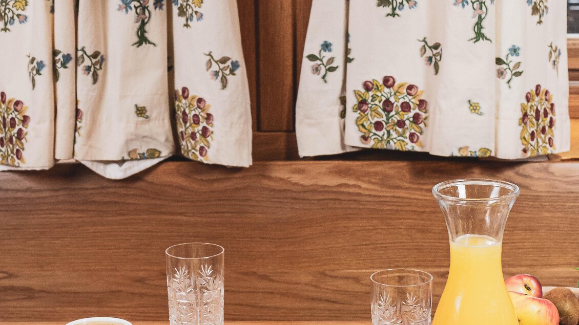 Ein gemütlicher Frühstückstisch mit verschiedenen Speisen wie Obst, Brot und Käse. Im Hintergrund hängen Vorhänge mit buntem Muster und einem Herz aus Stoff. | © Anna Ullrich