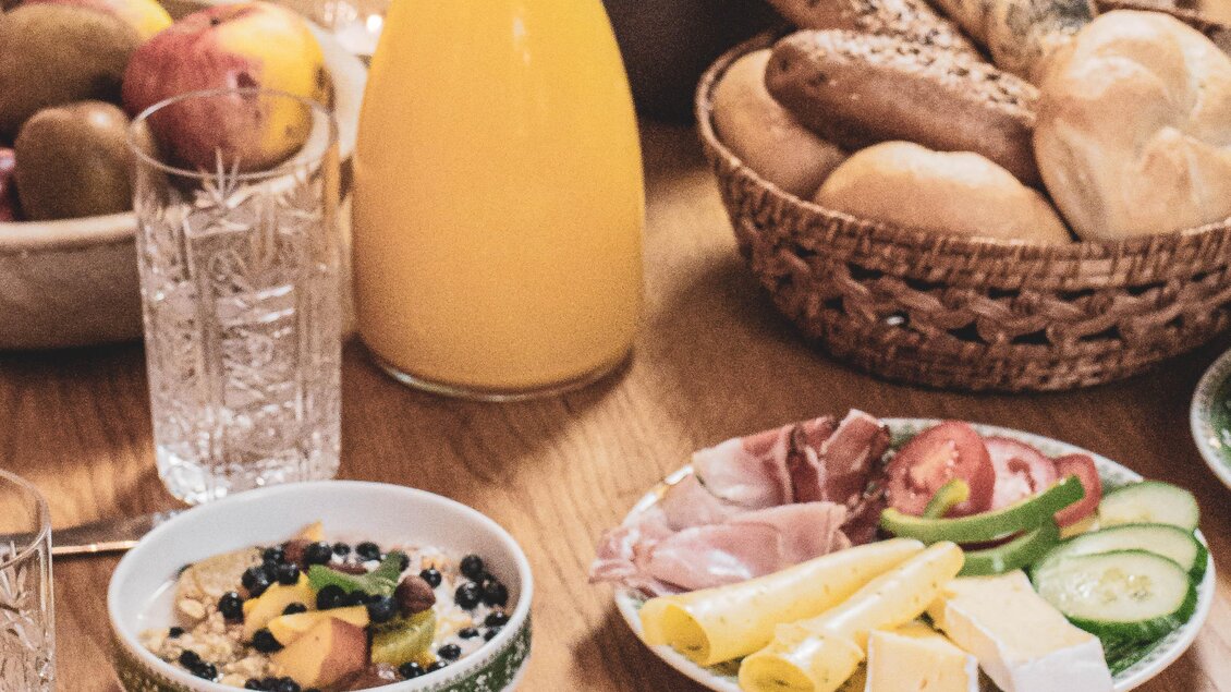 Ein reichhaltiges Frühstück mit verschiedenen Speisen steht auf einem Holztisch. Es gibt Brötchen, Käse, Wurst, frisches Obst und Getränke. | © Anna Ullrich