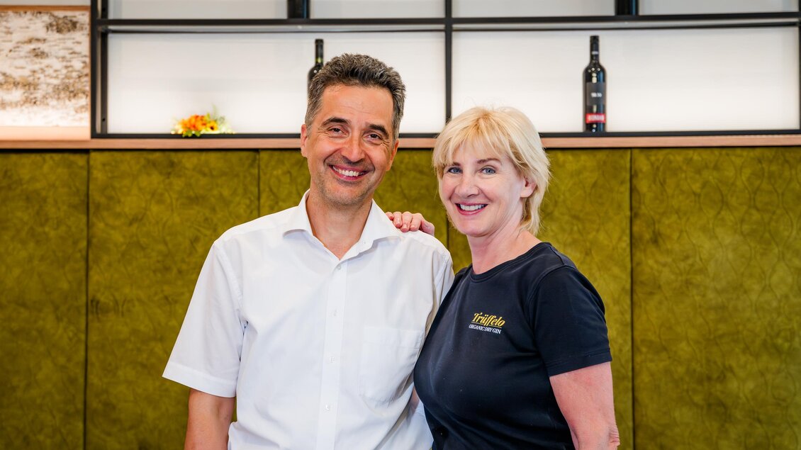 Ein freundliches Paar lächelt vor einer modern gestalteten Wand. Im Hintergrund stehen einige Weinflaschen. | © Region Graz - Mias Photoart