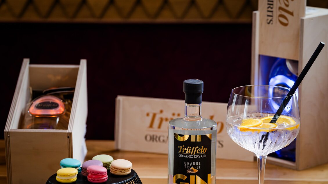 Ein stilvoller Tisch mit einer Flasche Gin, einem Cocktailglas mit Zitrone und bunten Macarons. Im Hintergrund sind elegante Holzkisten zu sehen. | © Region Graz - Mias Photoart