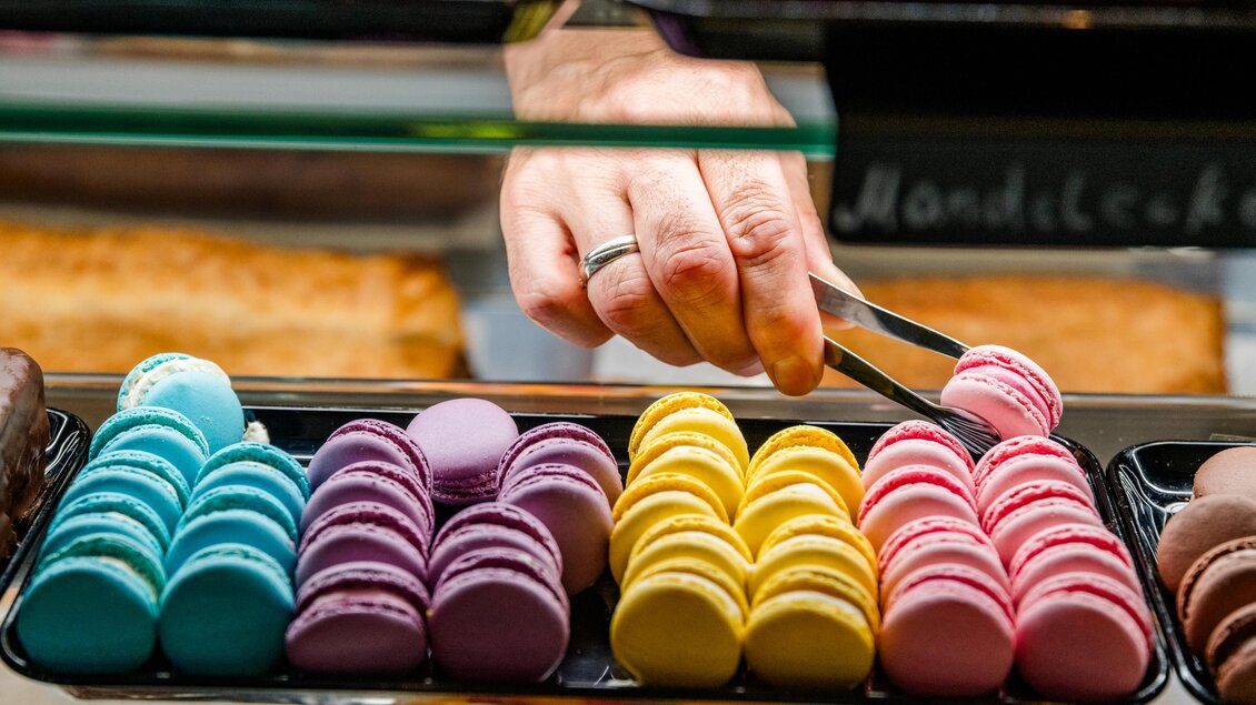 Bunte Macarons in verschiedenen Farben stehen auf einem Tablett. Eine Hand greift mit einer Zange nach einem Macaron. | © Region Graz - Mias Photoart