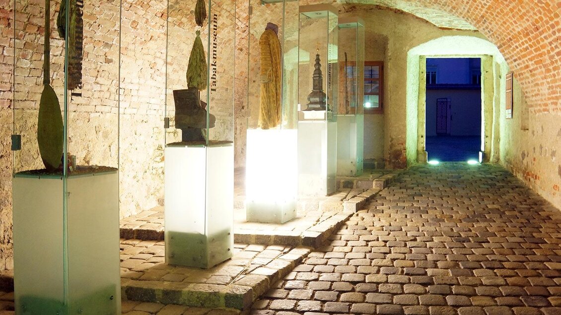 Ein historischer Korridor mit Steinboden und Ziegelwänden. Skulpturen in Glasvitrinen sind an den Seiten ausgestellt, während der Ausgang im Hintergrund beleuchtet ist. | © Uwe Reinecker