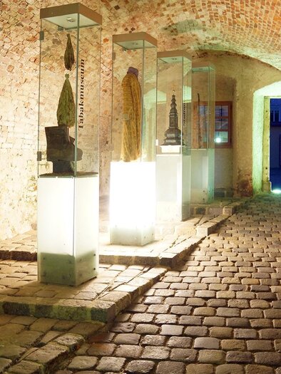 Ein historischer Korridor mit Steinboden und Ziegelwänden. Skulpturen in Glasvitrinen sind an den Seiten ausgestellt, während der Ausgang im Hintergrund beleuchtet ist. | © Uwe Reinecker