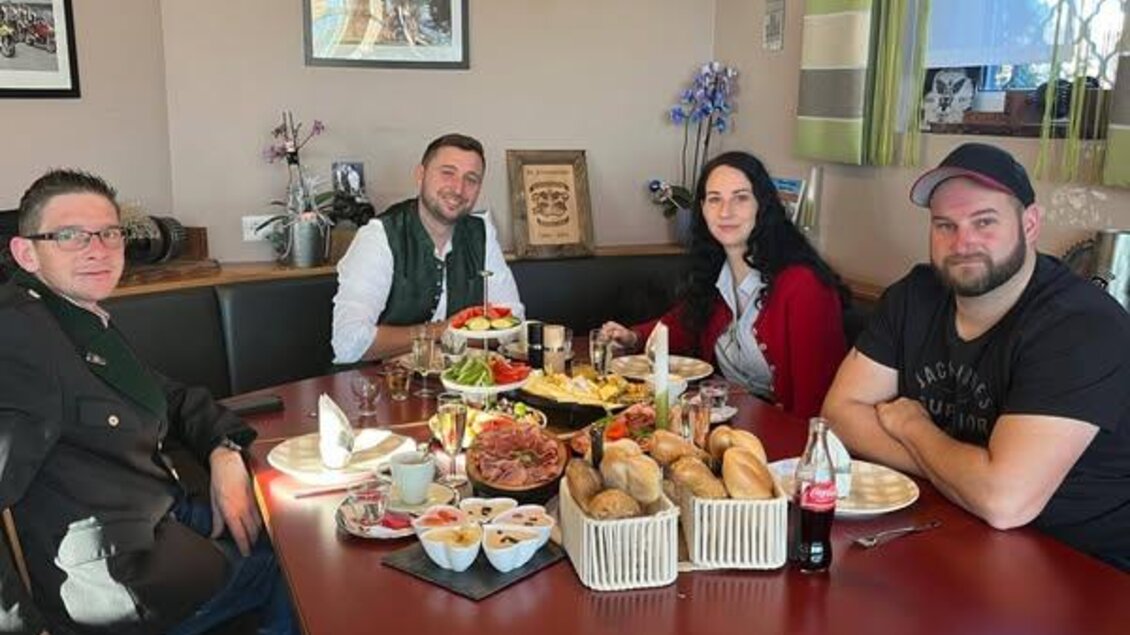 Eine Gruppe von vier Personen sitzt an einem Tisch, der mit verschiedenen Speisen und Getränken gedeckt ist. Im Hintergrund sind Bilder und eine grüne Gardine zu sehen. | © Bettina Fiedler