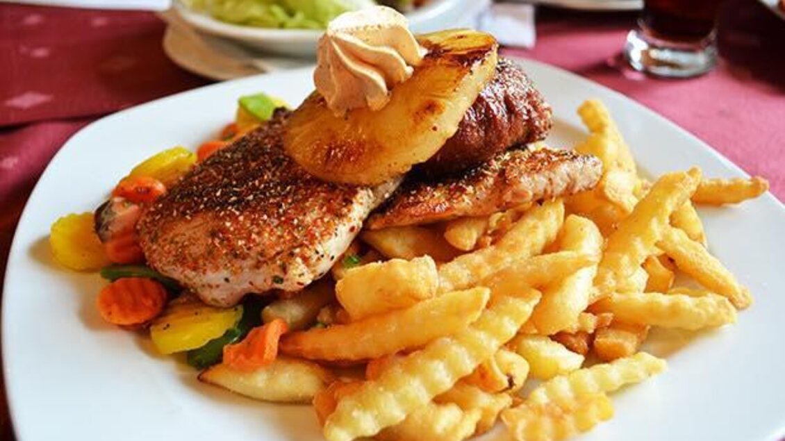 Ein Teller mit gebratenem Fischfilet, belegt mit Ananas und einer Sauce. Dazu gibt es gewellte Pommes frites und buntes Gemüse. | © Bettina Fiedler