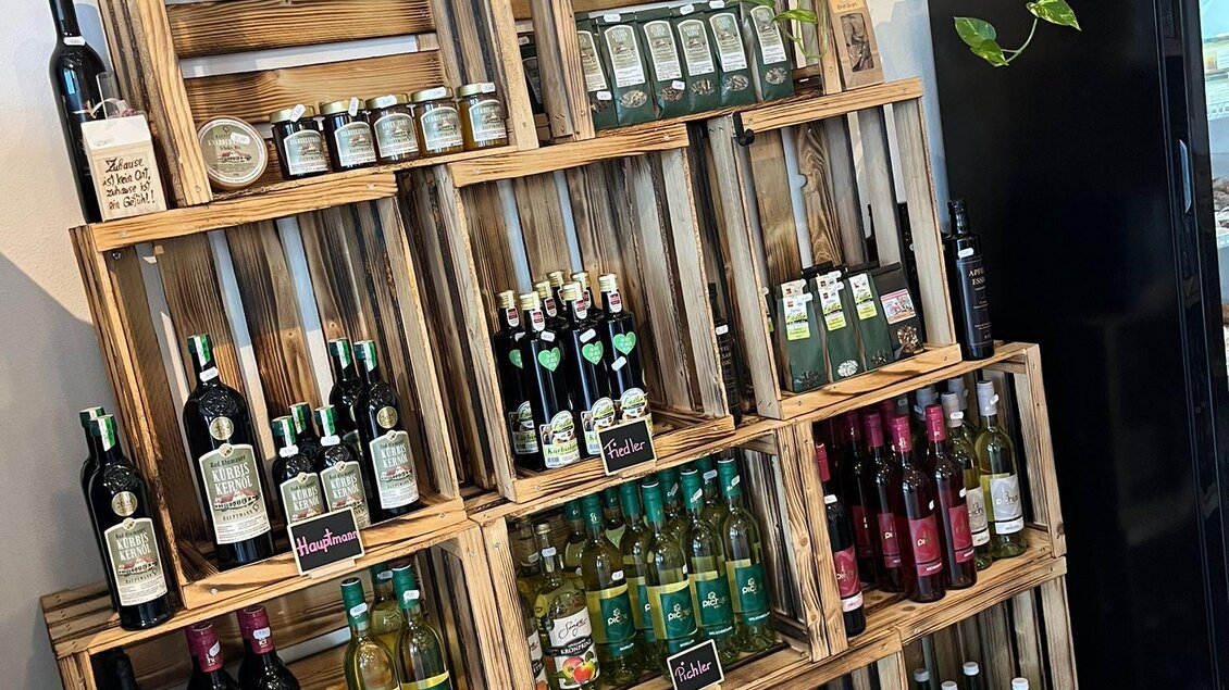 Ein Holzregal mit verschiedenen Flaschen, darunter Wein und Säfte. Die Flaschen sind in mehreren Ebenen angeordnet und einige Pflanzen sind dekorativ hinzugefügt. | © Bettina Fiedler