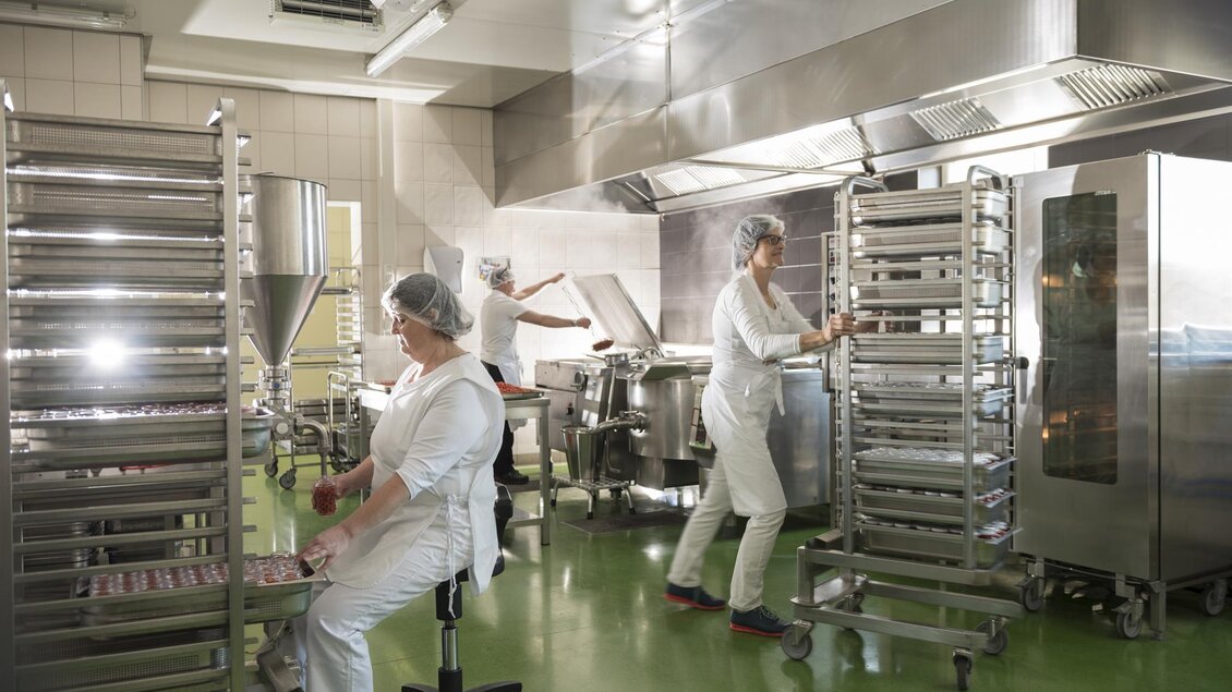 Eine moderne Küche mit mehreren Mitarbeitern, die an verschiedenen Aufgaben arbeiten. Es gibt mehrere Edelstahlgeräte und Regale in der Umgebung. | © Bernhard Bergmann