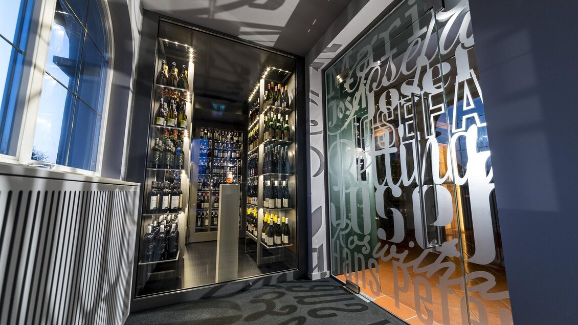 Ein modischer Eingangsbereich mit Glastüren und stilvollem Schriftzug. Im Hintergrund sind Regale mit Weinflaschen sichtbar. | © Werner Krug