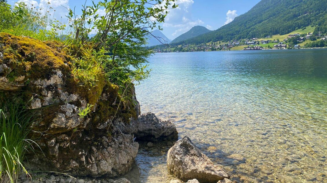 Ein klarer See umgeben von grünen Bergen und Bäumen. Felsen und Kies am Ufer spiegeln die ruhige Wasseroberfläche wider. | © Bettina Scheck