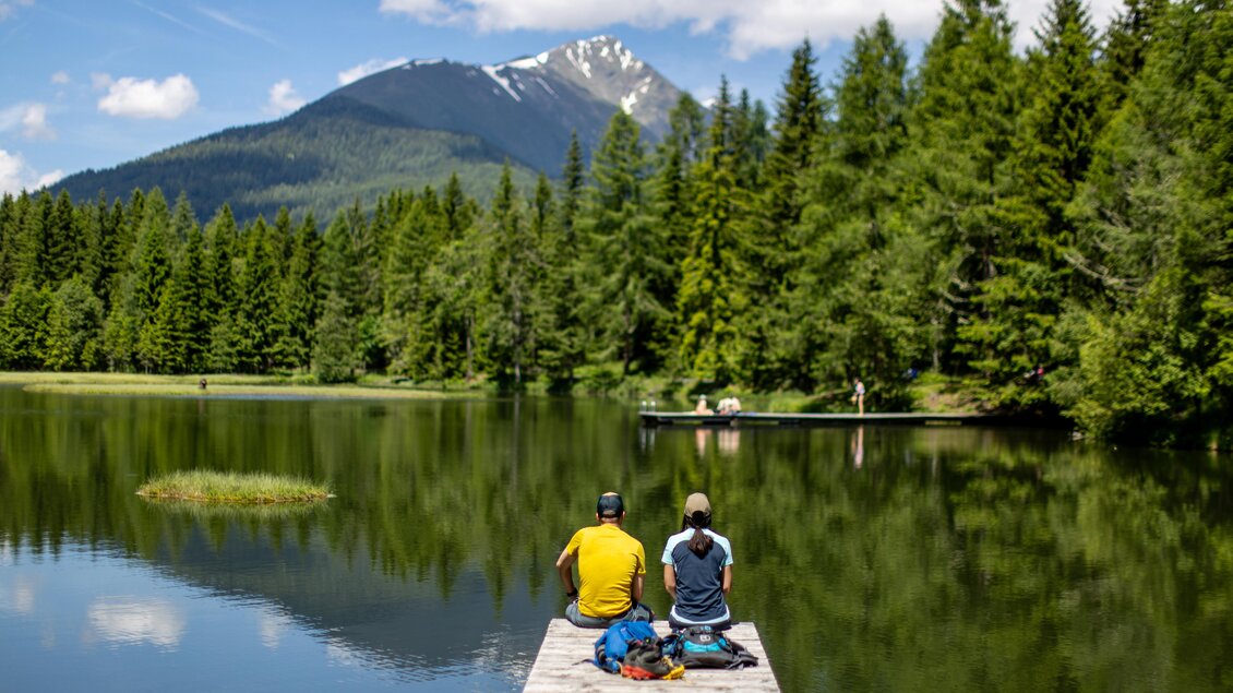Zwei Personen sitzen auf einem Bootssteg am Wasser. Im Hintergrund sind Berge und dichte Wälder zu sehen. | © TVB Murau