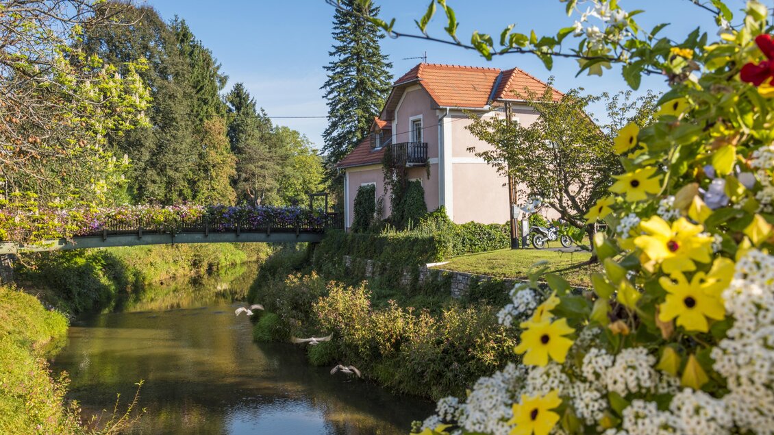 Ein malerisches Haus am Ufer eines ruhigen Flusses, umgeben von Bäumen und bunten Blumen. Im Hintergrund erstrahlt ein klarer blauer Himmel. | © Thermen- und Vulkanland