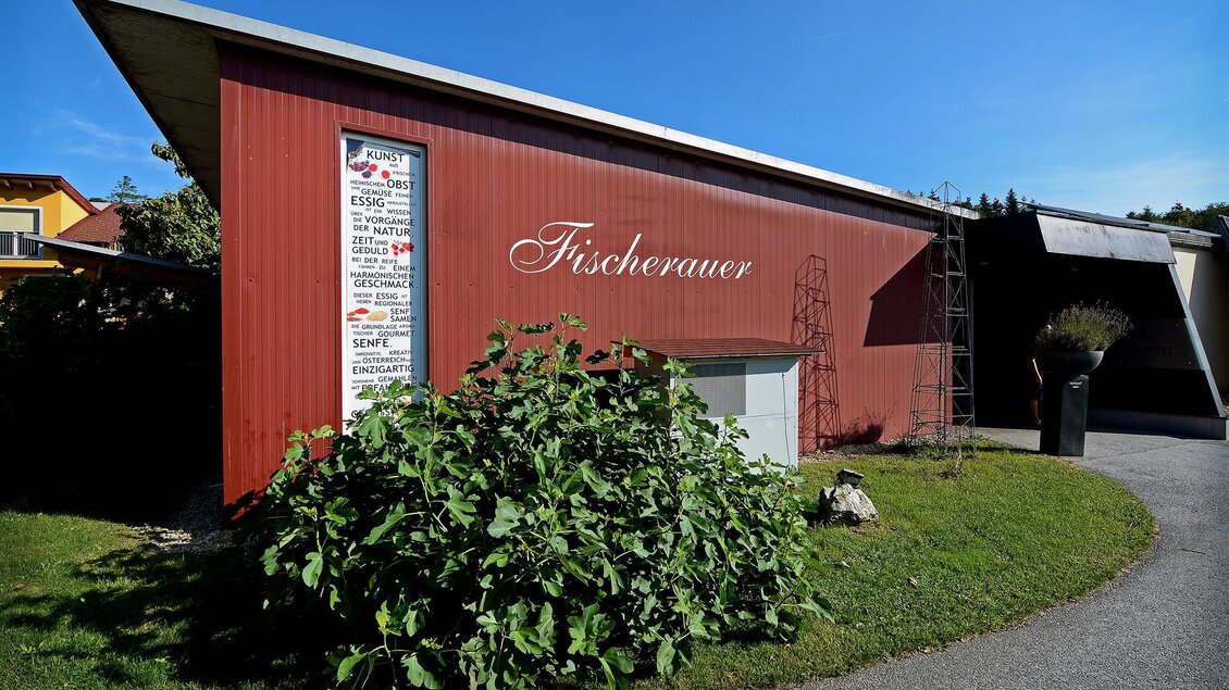 Ein rotes Gebäude mit dem Schriftzug "Fischerauer" in einer grünen Umgebung. Ein klarer blauer Himmel ergänzt die Szenerie. | © Tourismusverband Oststeiermark