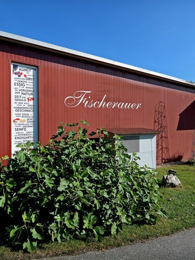 A red building with the inscription "Fischerauer" in a green environment. A clear blue sky complements the scene. | © Tourismusverband Oststeiermark