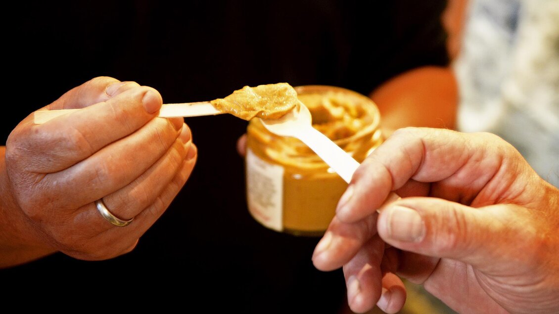 Ein Mann hält einen Löffel mit Erdnussbutter, während ein anderer Mann ihn anfasst. Die Szene zeigt die Interaktion zwischen den beiden Personen und die Erdnussbutter im Glas. | © Tourismusverband Oststeiermark