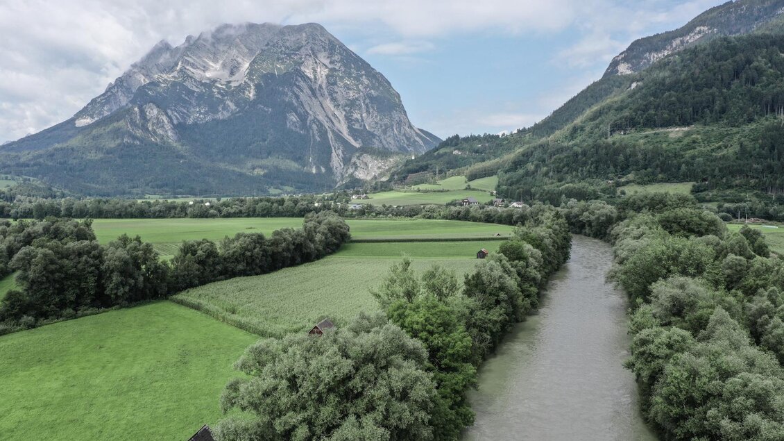 Eine malerische Landschaft mit einem Fluss, der durch grüne Wiesen fließt. Im Hintergrund erhebt sich ein beeindruckender Berg unter einem teilweise bewölkten Himmel. | © Fischereiverein Irdning