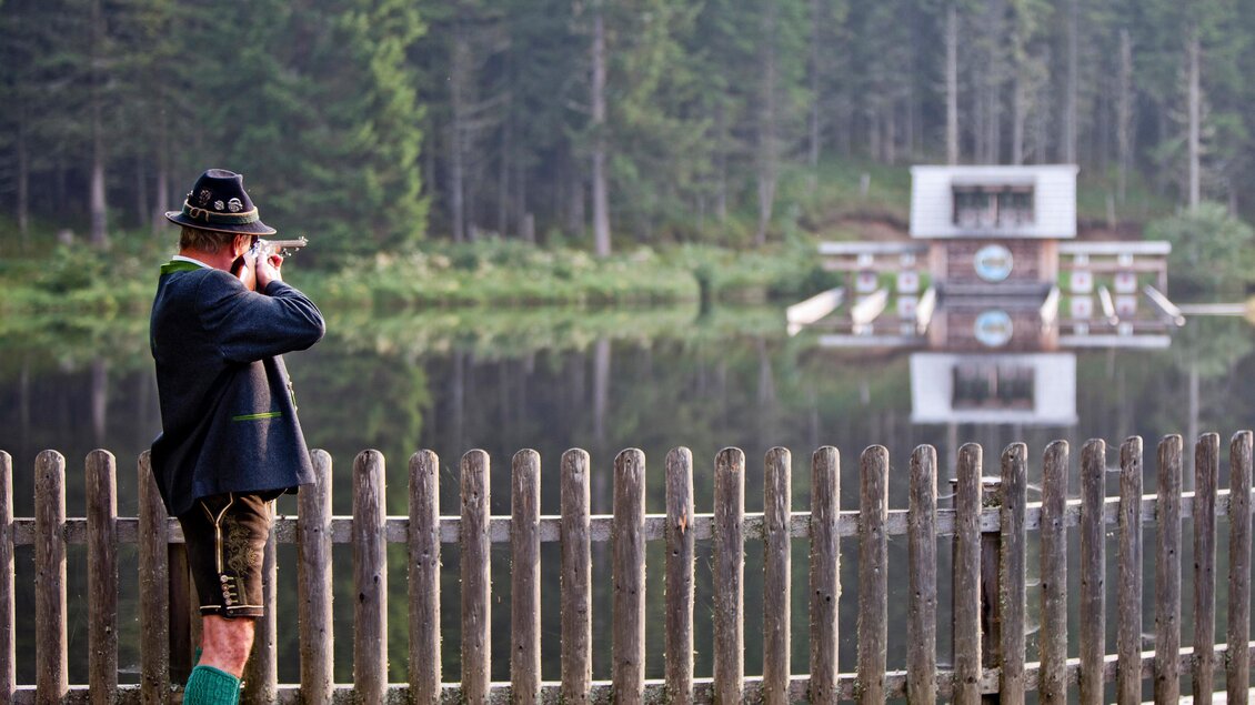 Ein Mann im traditionellen Outfit steht am Ufer eines ruhigen Sees und blickt durch ein Gewehr. Im Hintergrund sind Bäume und ein Gebäude zu sehen, das sich im Wasser spiegelt. | © Holzwelt Murau