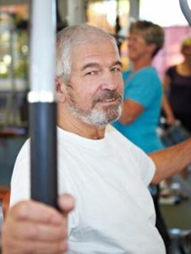 An older man is training at a strength machine in the gym. In the background, other people can be seen working out. | © Fotolia