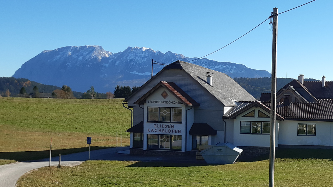 Ein schönes Gebäude mit einem Schild für Kachelöfen steht vor einer malerischen Berglandschaft. Im Hintergrund sieht man schneebedeckte Berge unter einem klaren blauen Himmel. | © TVB Ausseerland Salzkammergut_Daniela Casari