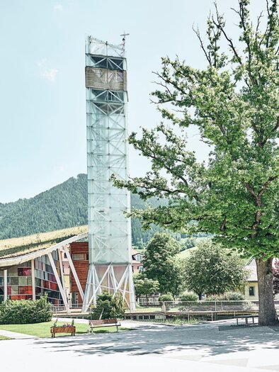 A modern building with a glass tower stands in a quiet environment. Gentle hills and trees can be seen in the background. | © Armin Walcher
