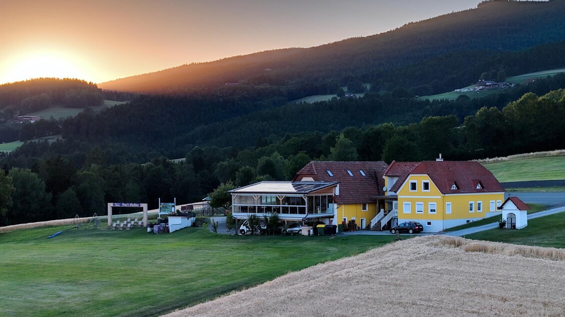 Ein schönes Landhaus inmitten sanfter Hügel. Die Sonne geht hinter den Bergen unter und taucht die Landschaft in warmes Licht. | © Flourl´s Schenke
