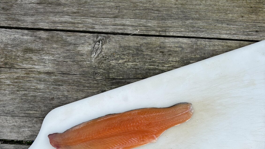 Ein frischer Fischfilet liegt auf einem weißen Schneidebrett. Im Hintergrund sind einige Küchenutensilien und ein Holzuntergrund sichtbar. | © Selina Senft