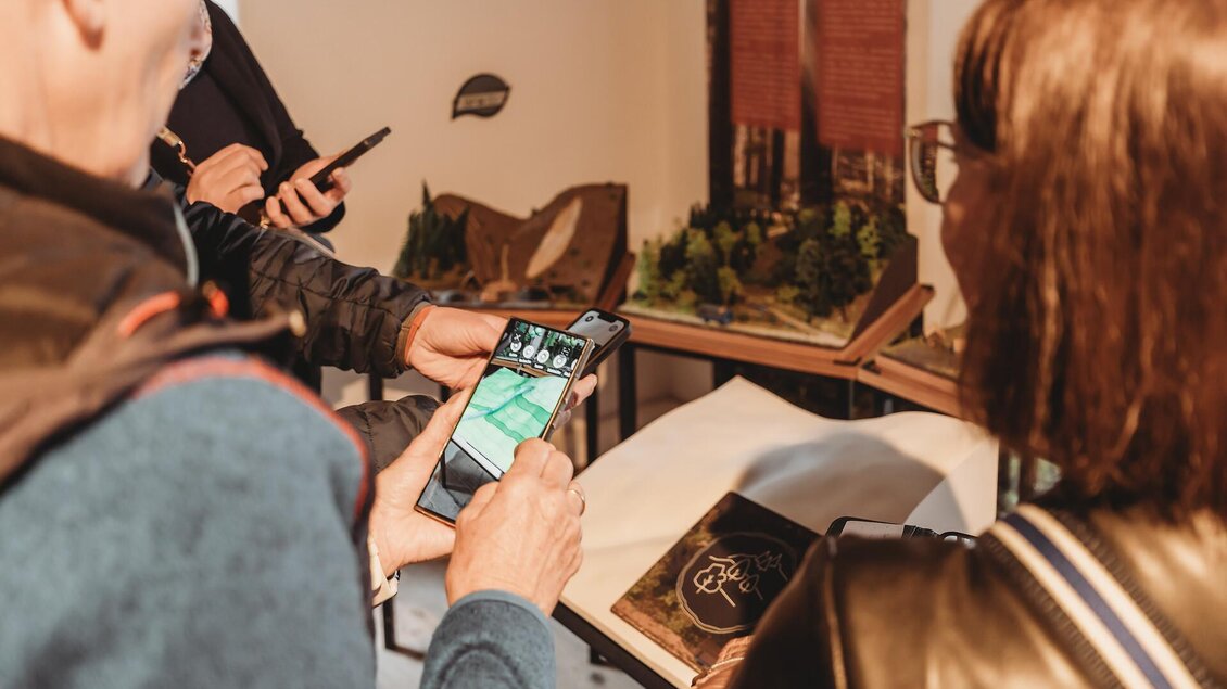 Eine Gruppe von Personen betrachtet ein Smartphone, während sie an einem Tisch stehen. Auf dem Tisch sind Modelle und andere Objekte zu sehen.