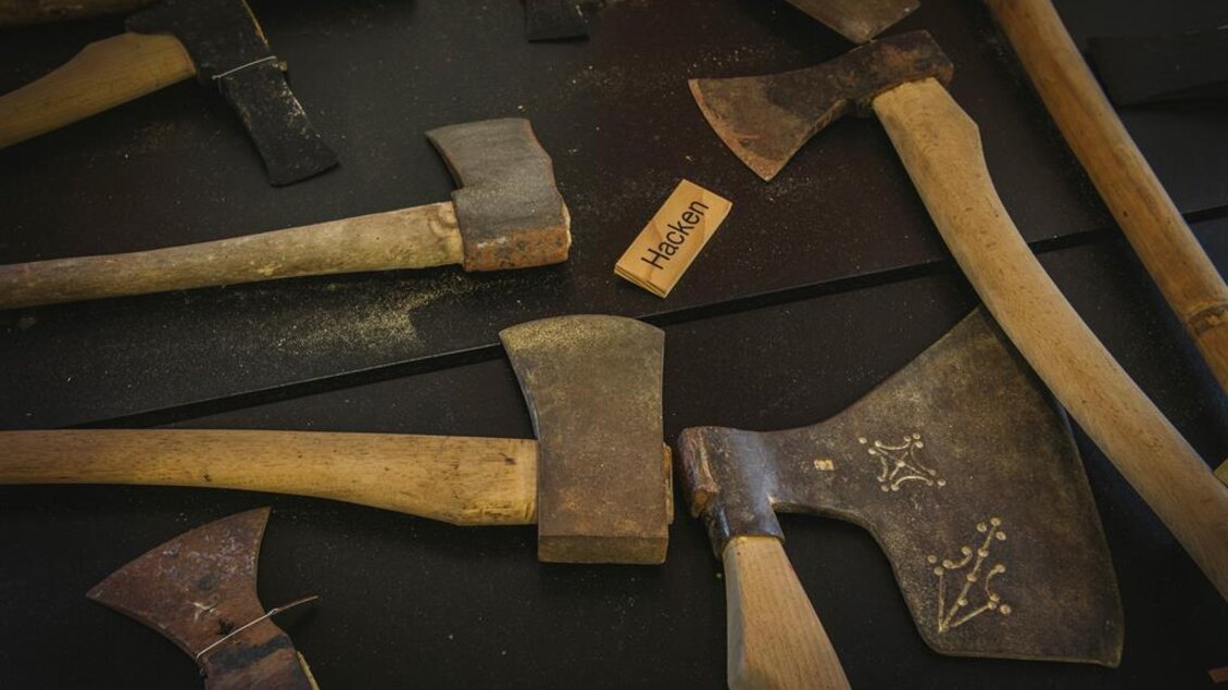 Eine Sammlung von Äxten mit hölzernen Griffen auf einem dunklen Hintergrund. Eine Beschriftung „Hacken“ ist ebenfalls sichtbar. | © Stefan Leitner