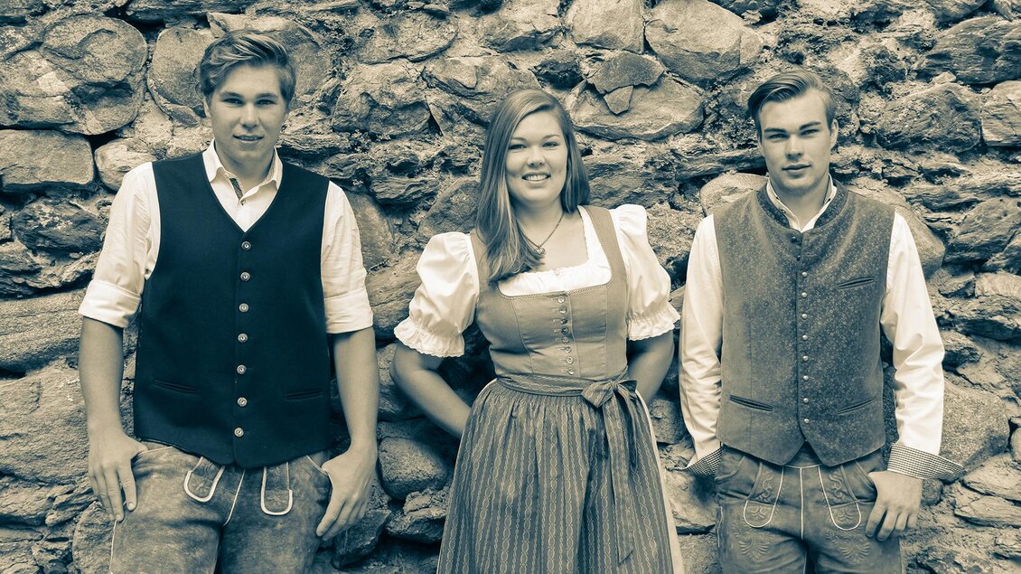 Drei Personen in traditioneller Kleidung stehen vor einer Steinmauer. Sie lächeln und wirken freundlich.