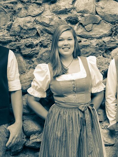 Three people in traditional clothing stand in front of a stone wall. They are smiling and appear friendly.