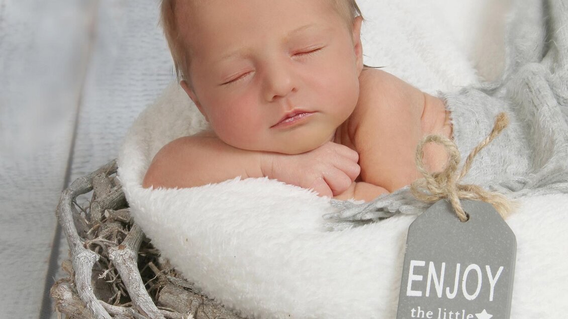 Ein schlafendes Baby in einer gemütlichen Umgebung mit einem weichen Tuch. Ein Schild mit der Aufschrift „Enjoy the little things“ liegt daneben.