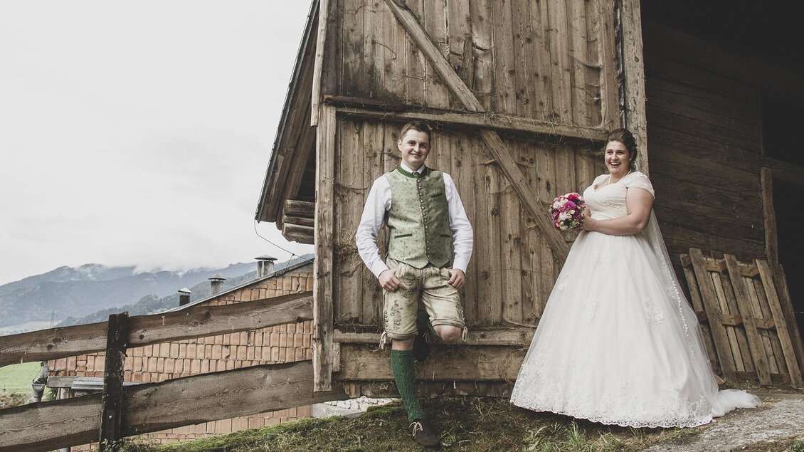 Ein Brautpaar steht vor einer Scheune. Die Braut trägt ein weißes Kleid und der Bräutigam trägt eine Tracht.