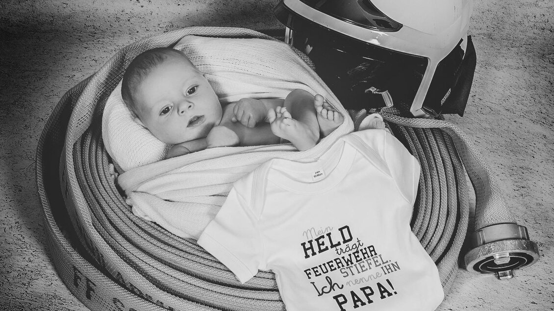 Ein Baby liegt in einer Decke, umgeben von Feuerwehrschlauch und einem Feuerwehrhelm. Es trägt einen weißen Body mit der Aufschrift „Held, ich bin der Papa!“