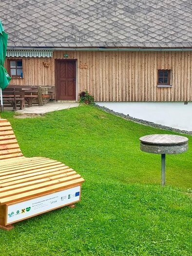 Eine gemütliche Liege aus Holz steht auf einer grünen Wiese. Im Hintergrund sind Holzbungalows und ein Sonnenschirm zu sehen. | © Erlebnisregion Murtal