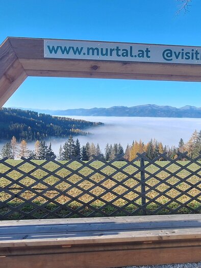 A wooden frame with a view of a picturesque landscape. In the background, fog and mountains are visible. | © Erlebnisregion Murtal