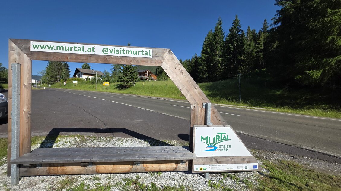 Ein Holzschild mit der Aufschrift "www.murtal.at" und "visitmurtal", das Besucher anzieht. Im Hintergrund sind grüne Bäume und eine klare Straße zu sehen. | © Marlene Zandl