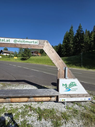 A wooden sign with the inscription "www.murtal.at" and "visitmurtal" that attracts visitors. In the background, there are green trees and a clear road visible. | © Marlene Zandl