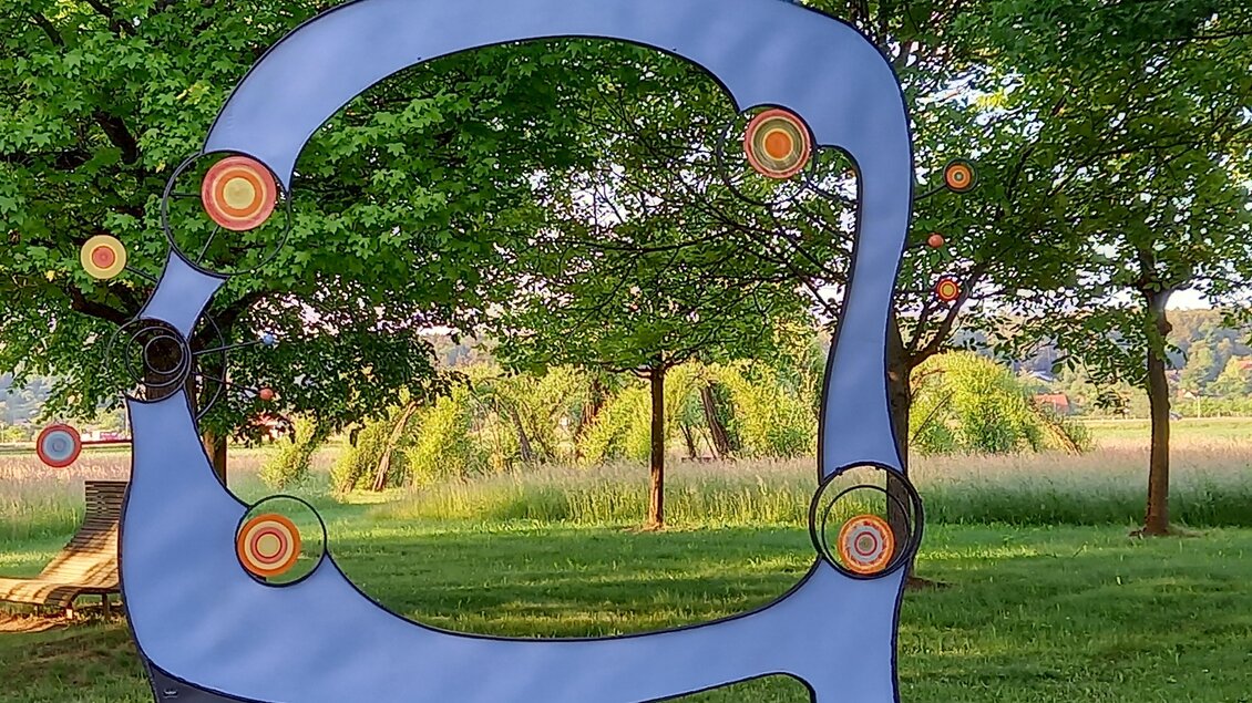 Eine moderne Skulptur in einem grünen Park. Sie hat eine geschwungene Form und farbige Kreise, die an eine Zielscheibe erinnern. | © Thermen- & Vulkanland