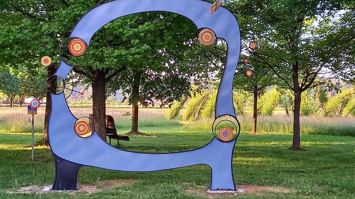 Eine moderne Skulptur aus buntem Metall steht in einem Park. Umgeben von Bäumen und grünem Gras, zeigt sie verschiedene Kreise und Formen. | © Thermen- & Vulkanland