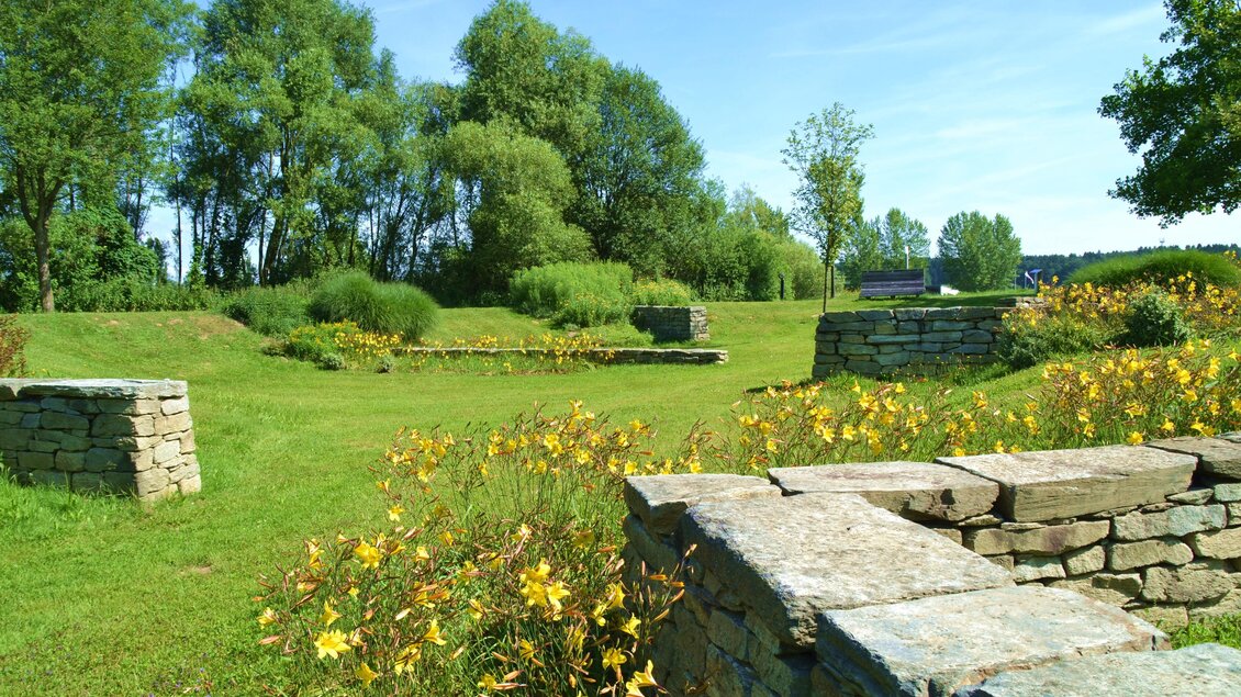 Eine große Wiese mit bunten Blumen und niedrigen Steinmauern. Im Hintergrund sind Bäume und ein klarer blauer Himmel sichtbar. | © Kurkommission Bad Blumau