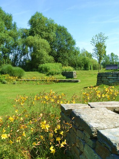 A large meadow with colorful flowers and low stone walls. In the background, trees and a clear blue sky are visible. | © Kurkommission Bad Blumau