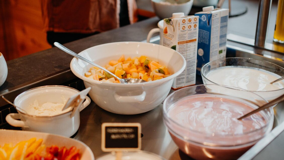 Ein reichhaltiges Frühstücksbuffet mit verschiedenen Schüsseln, darunter Joghurt, Obst und Salat. Im Hintergrund ist eine Person zu sehen, die sich am Buffet bedient.