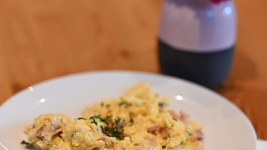 Ein Teller mit Rührei, garniert mit Kräutern und Schinkenstücken. Im Hintergrund steht eine kleine Vase mit einer Blume.