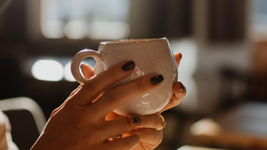 Eine Hand hält eine weiße Tasse in einem sonnigen Café. Der Hintergrund zeigt verschwommene Fenster und eine warme Atmosphäre.